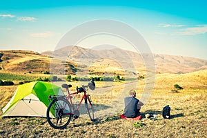 Caucasian man sit by touring bicycle in mountains on side of green tent have break time rest in morning. Solo travel journey. Long