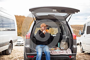 Caucasian man sits in the trunk of a car and drinks coffee. Outdoor picnic. Road trip