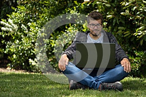 Caucasian man sitting on the grass of a park and looking at the screen of his laptop.