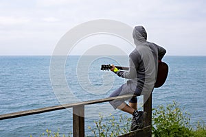 Caucasian man sitting on railing at the coast of the sea and playing guitar. view from the back