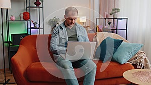 Caucasian man sitting on sofa opening laptop pc starting work e-learning online in room at home