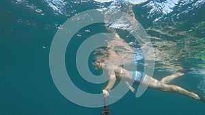 A Caucasian man in a snorkel mask and flippers dives and swims underwater with an action camera on a long selfie stick