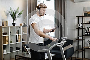 Caucasian man in sport clothes using headset working tablet computer while doing cardio training