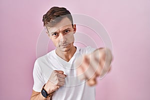 Caucasian man standing over pink background punching fist to fight, aggressive and angry attack, threat and violence