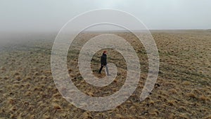 A caucasian man in tourist clothing walks on the top of a plateau next to a deep cliff along with a drone in fog and low