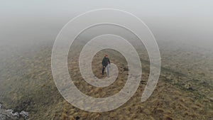 A caucasian man in tourist clothing walks on the top of a plateau next to a deep cliff along with a drone in fog and low