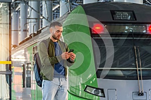 Caucasian man traveler in green windbreaker and with backpack stands on rail road platform typing text on phone, electric commuter
