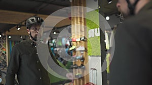 Caucasian man trying on black bicycle helmet near mirror in sporting goods store. Male buyer chooses safety helmet for