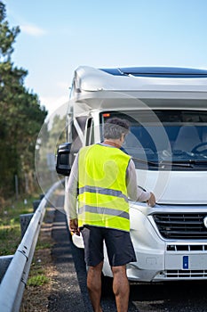 Caucasian man waiting a mechanic to come fix his broken down motor home on the highway with a reflective vest and checking the