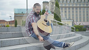 Caucasian man walking up urban stairs, taking off hat, and start playing guitar. Portrait of street musician performing