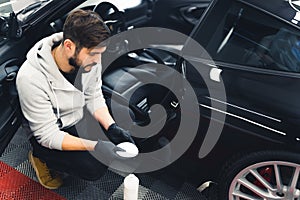 Caucasian man wearing black gloves squatting next to car preparing leather proofing liquid to put on car seat upholstery