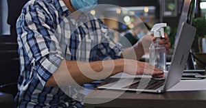 Caucasian man wearing face mask cleaning his laptop using tissue and disinfectant spray at modern of