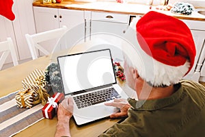 Caucasian man wearing santa hat, sitting at table in kitchen, using laptop with copyspace