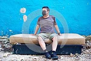 Caucasian man wears medical mask and sits on a discarded bed. Coronavirus and homeless protection