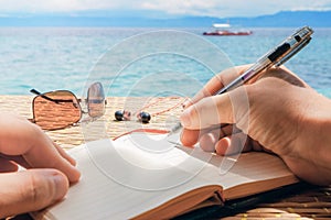 Caucasian man is writing sime idea, message or letter in his notepad by pen while he sitting on the beach of tropical sea with boa