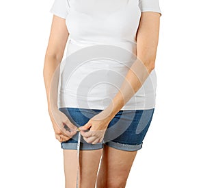 Caucasian mature adult woman begins to gain weight and measures her thigh with a measuring tape. Woman dressed in a white T-shirt