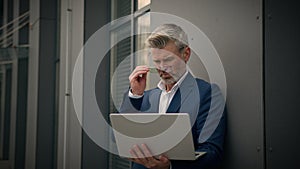 Caucasian mature retired business man with laptop working distant online work in city leaning to wall off company office