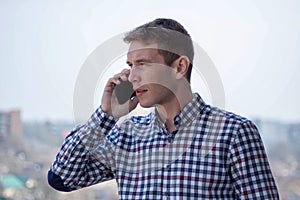 Caucasian  men  is making a business call. There is a city on the back ground.  Dressed into checkered shirt. Stands on the 9th photo