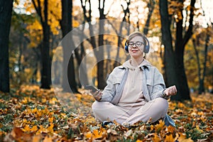 Caucasian middle aged woman in earphones listening to music, meditation app on smartphone and meditating in lotus pose