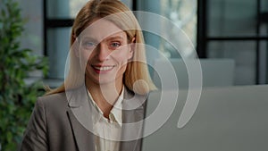 Caucasian middle-aged woman in office with computer working online smiling to camera. Business portrait female