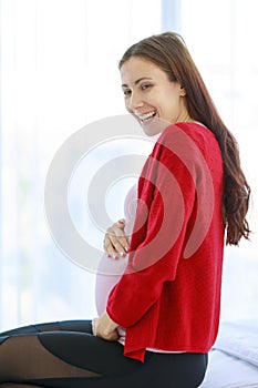 Caucasian millennial young happy female prenatal pregnant mother model in casual pregnancy outfit jacket sitting alone on bed in