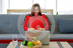 Caucasian millennial young happy sexy female prenatal pregnant mother in casual red pregnancy dress sitting on sofa holding hands