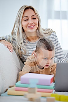 caucasian mom and little boy sitting on sofa at home watch video on modern laptop
