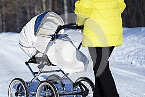 Caucasian mom in yellow coat is walking with white baby carriage on snow road among the forest at winter sunny day