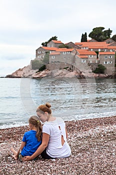 Caucasian mother and daughter sitting together on the luxury beach against Sveti Stefan hotel and resort. Adriatic sea, Montenegro