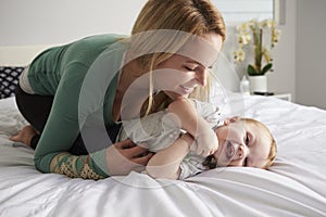 Caucasian mother kneeling on bed, tickling her baby daughter