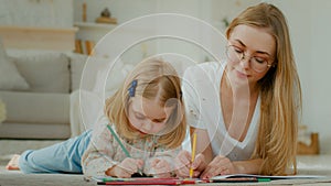 Caucasian mother lying of floor at home with little small adopted child girl enjoy draw together. Daughter baby