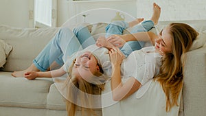 Caucasian mother mom with daughter lying on couch sofa in living room mommy tickle kid girl laughing tickling child baby