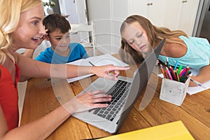 Caucasian mother using laptop and doing homework with her daughter and son smiling at home