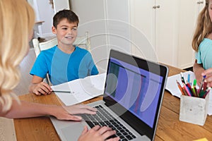 Caucasian mother using laptop and doing homework with her daughter and son smiling in kitchen