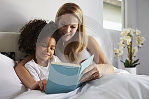 Caucasian mum and black daughter read a book together in bed