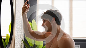 Caucasian naked man guy male in bathroom morning beauty routine procedure looking at mirror reflection apply spray hair