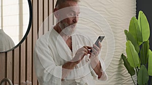 Caucasian old retired man mature senior male in bathrobe morning home hotel bathroom reading smartphone message mobile