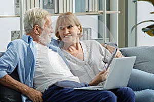 Caucasian old senior elderly grandparents couple sitting cuddling hugging talking together on sofa in living room at home. Gray