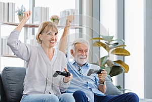 Caucasian old senior elderly lovely happy grandparents married couple husband and wife sitting hold fists up with joystick playing