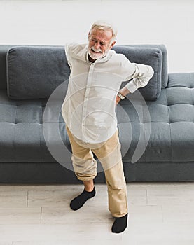 Caucasian old senior elderly unhealthy sick gray hair and bearded man standing near sofa in living room at home alone holding