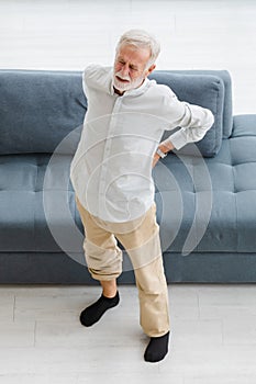 Caucasian old senior elderly unhealthy sick gray hair and bearded man standing near sofa in living room at home alone holding