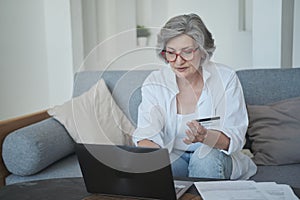 Caucasian older woman in eyewear using a credit card and a laptop to purchase online.