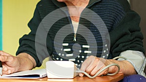 Caucasian Pensioner sitting at the table at home measures blood pressure and writes down the readings in a notebook, keeps statist