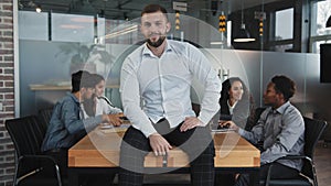 Caucasian pensive young male leader sitting on table in conference room confident boss smiling looking at camera