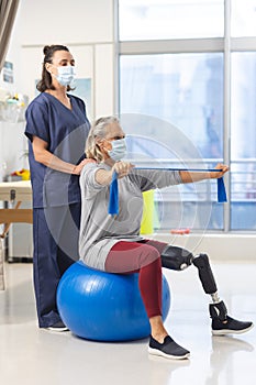 Caucasian physiotherapist and senior woman with artificial leg with face masks using exercise band