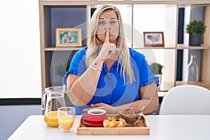 Caucasian plus size woman eating breakfast at home asking to be quiet with finger on lips