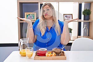 Caucasian plus size woman eating breakfast at home clueless and confused expression with arms and hands raised