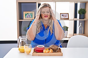 Caucasian plus size woman eating breakfast at home with hand on head for pain in head because stress