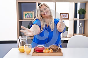 Caucasian plus size woman eating breakfast at home looking at the camera smiling with open arms for hug