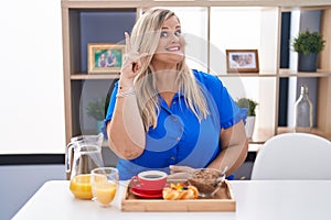 Caucasian plus size woman eating breakfast at home pointing finger up with successful idea